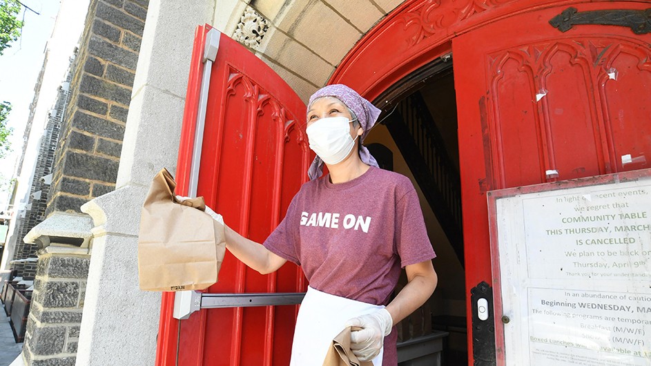 A woman with a bandana and a mask stands in front of an open red door, offering a bag of food. 