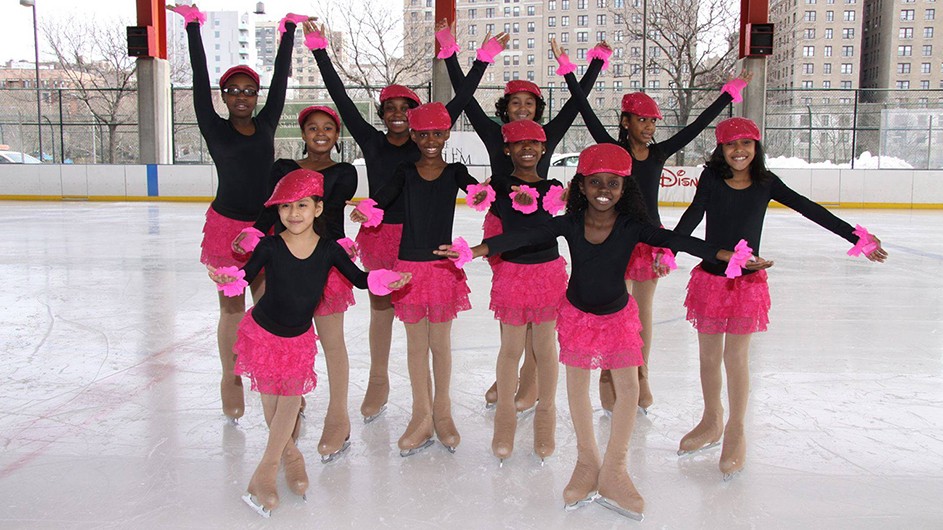 A group of kids posing on an ice rink in pink skirts and black leotards on skates. 