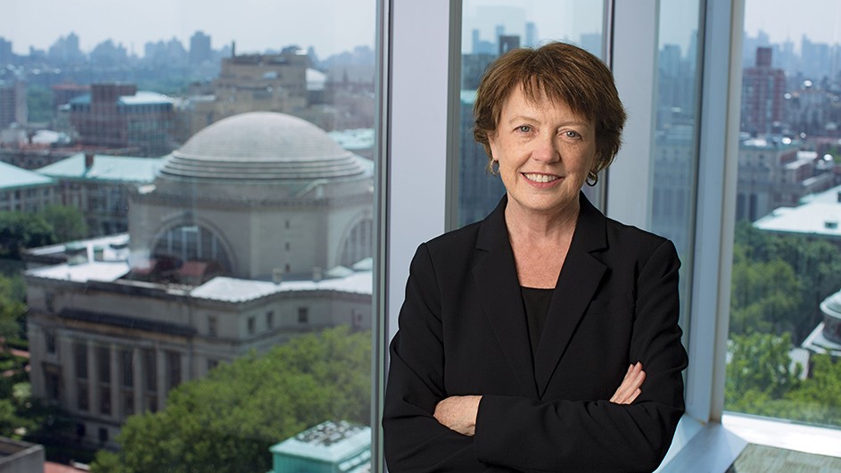 Jeffrey Schifman/Columbia Engineering Photo of a woman with short, medium-brown hair, in a dark suit, standing against a window overlooking Columbia's Morningside campus.