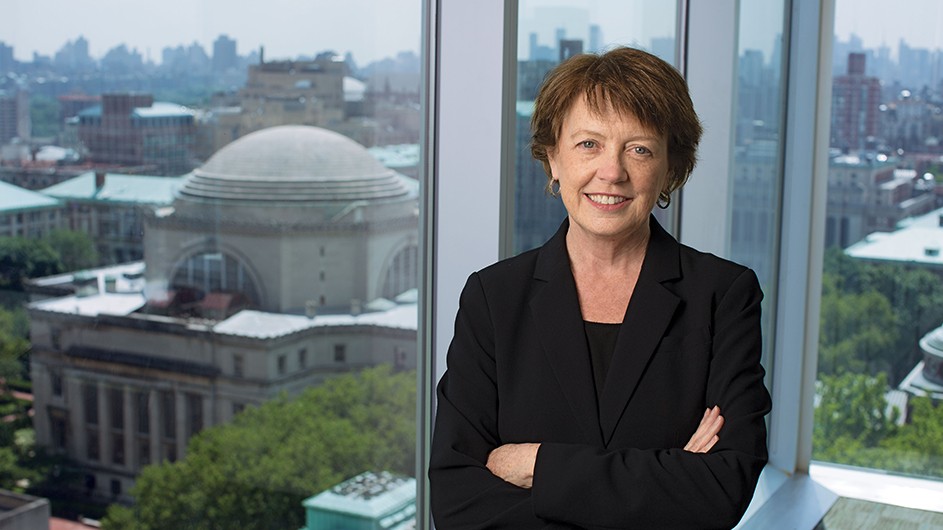 Photo of a woman with short, medium-brown hair, in a dark suit, standing against a window overlooking Columbia's Morningside campus.