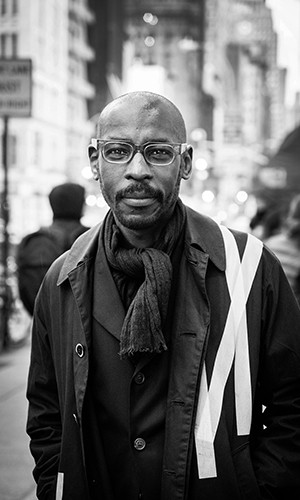 Black and white photo of Mario Gooden, a man with glasses in a dark shirt and jacket, against the backdrop of a cityscape. 