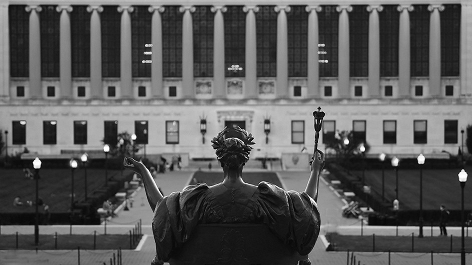Black and white photo of Alma Mater, a bronze statue of a woman sitting on a throne with a scepter and a laurel crown, viewed from the back. 