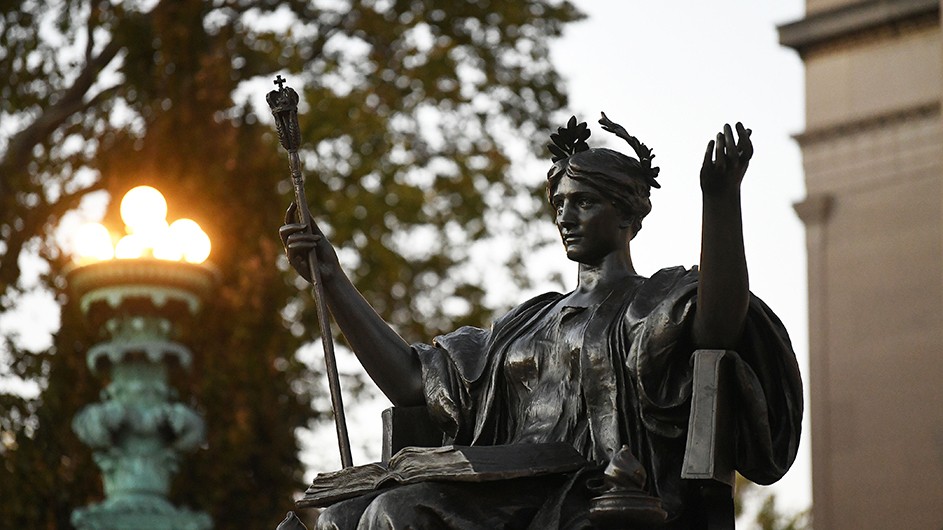 Side view, with warm lights, of Alma Mater, a bronze sculpture of a woman sitting down with a laurel wreath on her head and a scepter in her hand. 