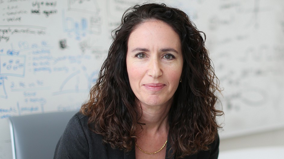 A woman with long, dark hair, against a whiteboard with writing on it.