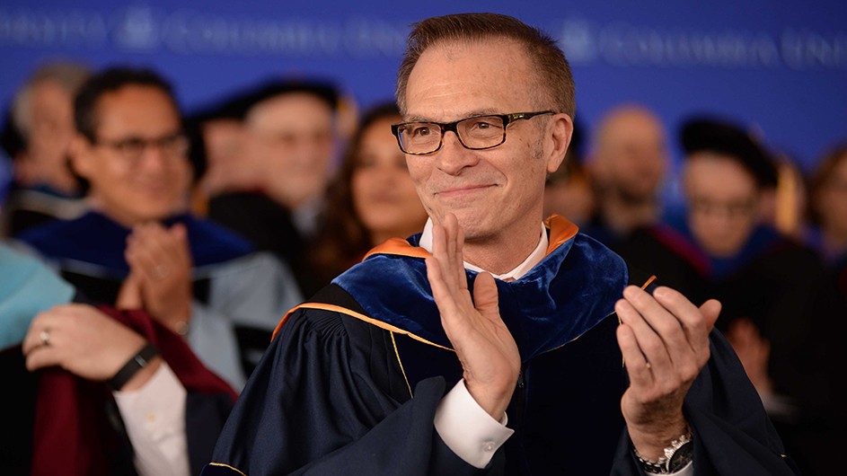 A man with short hair and glasses, sitting in an academic robe, clapping. 