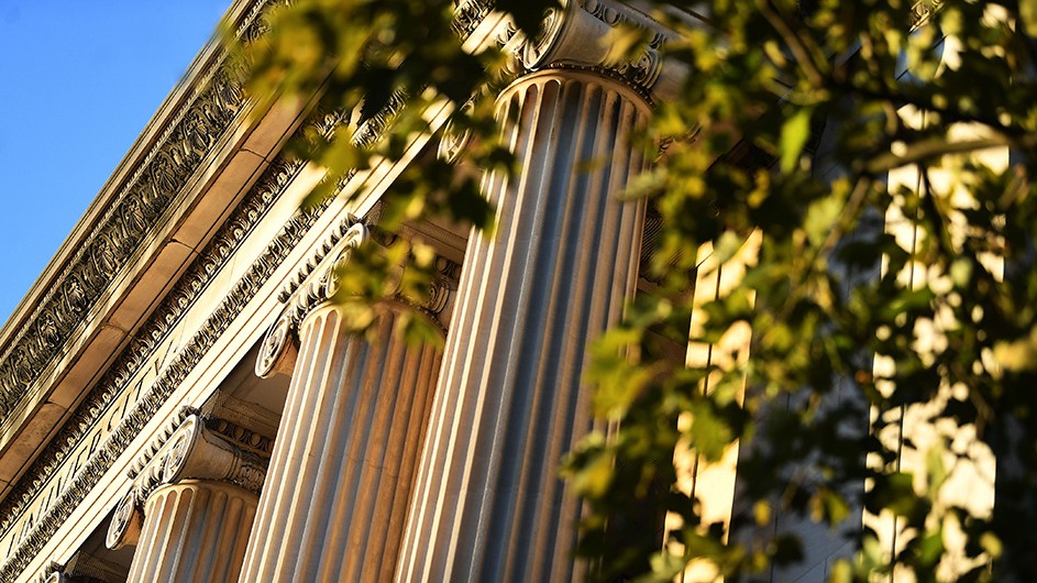 A view of the classical columns of Low Library on Columbia's Morningside campus, partially obscured by some green and leafy tree branches. 