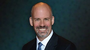 Man in a dark suit and tie, against a dark blue backdrop