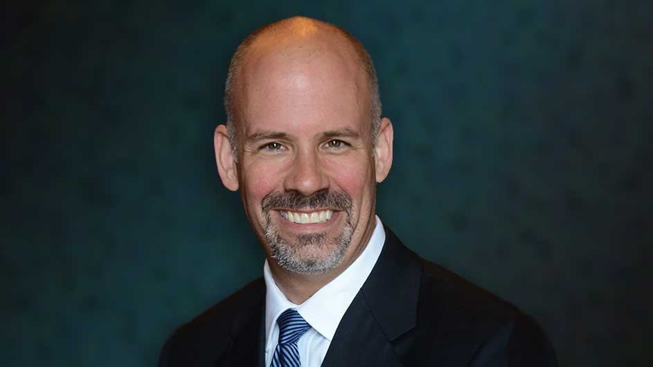 Man with mustache and beard in a dark suit, white shirt, and blue tie. 