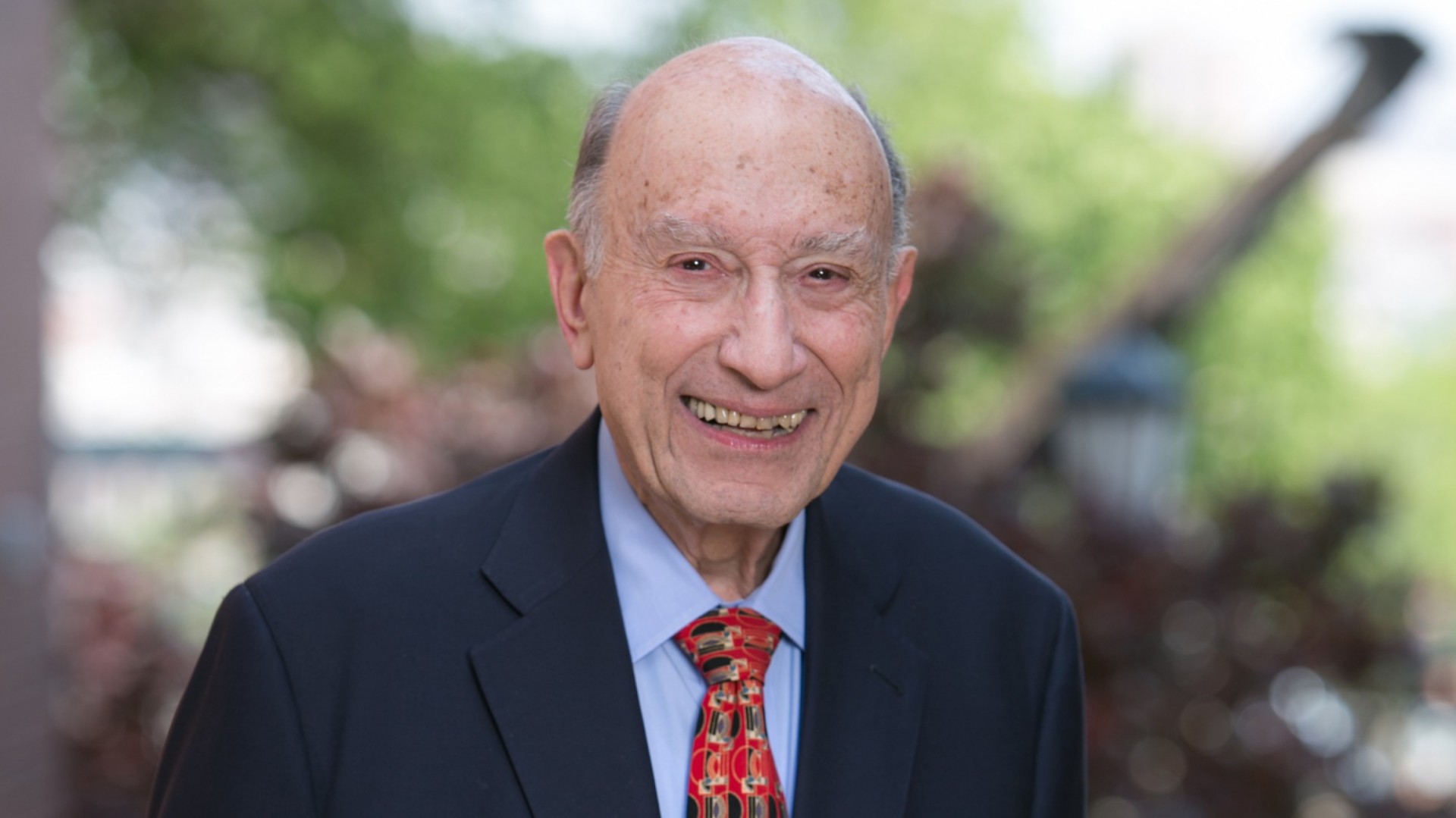 Head shot of Michael Sovern, older man with thinning hair, wearing a dark suit, blue shirt and red patterned tie.