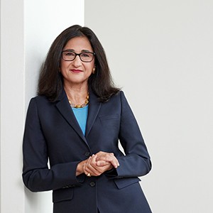 Minouche Shafik, a woman with long dark hair and glasses, smiling. 
