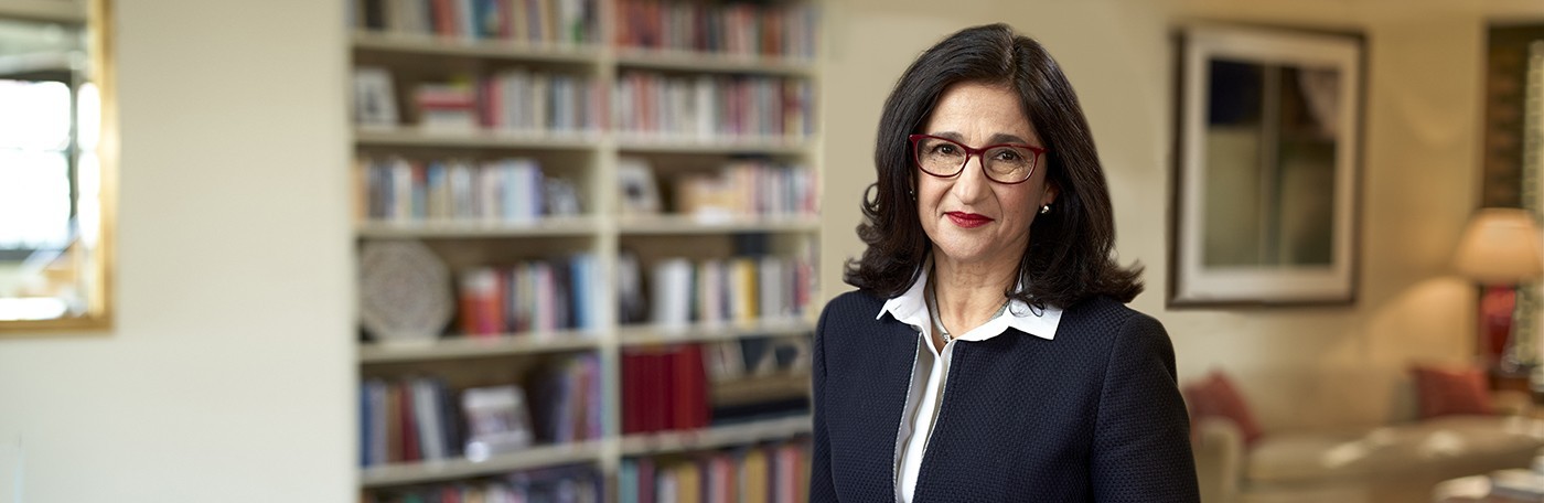 Wide image of a Minouche Shafik, a woman with long dark hair and glasses, smiling.