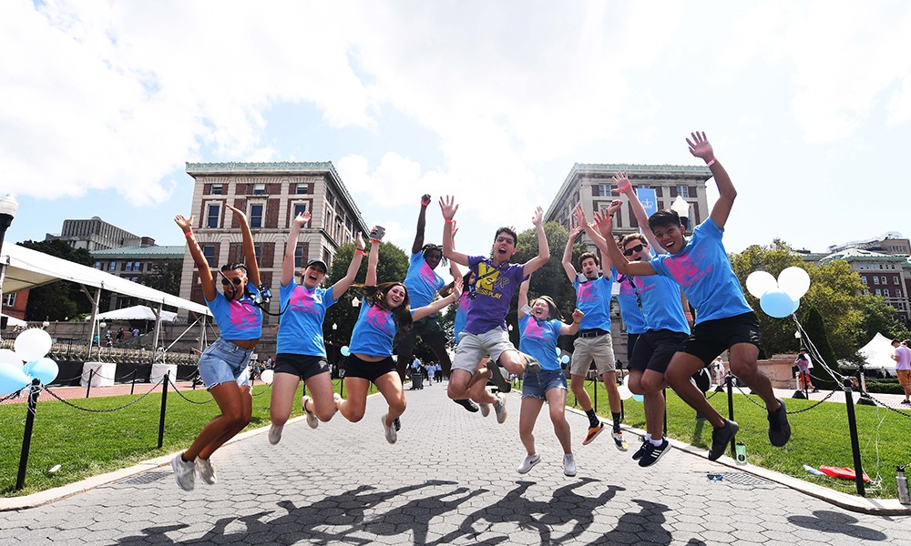 Photo of a group of students in blue shirts jumping in unison on college walk. 