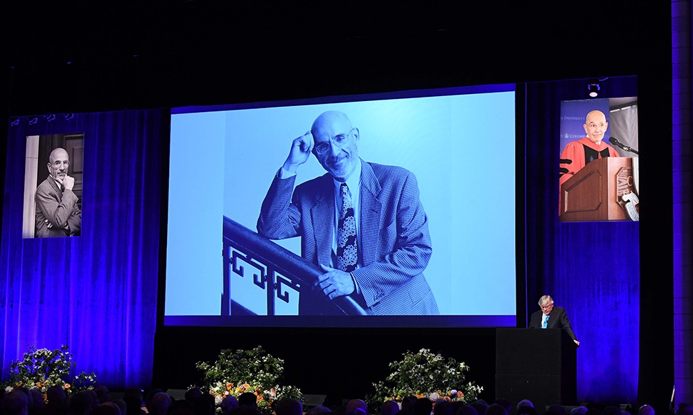Photo of President Bollinger speaking at Peter Awn's memorial, in front of three screens with pictures of Peter Awn on them.