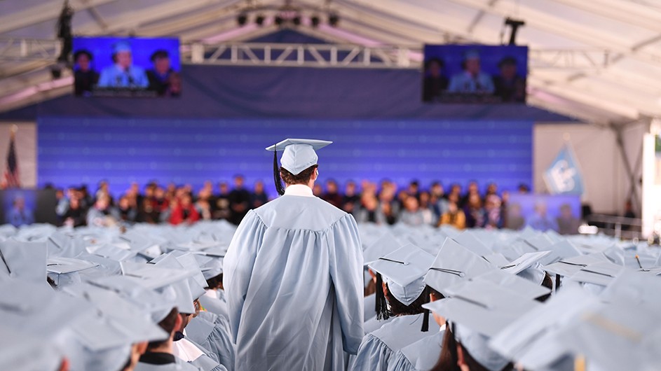 A student walks down an aisle at a graduation ceremony, surrounded by a sea of blue caps and gowns. 