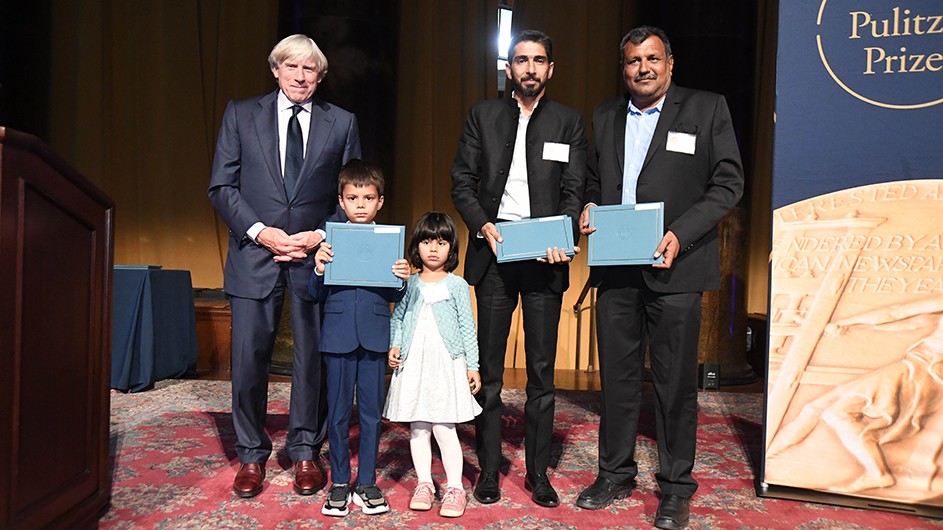 Columbia University President Lee C. Bollinger presents the 2022 Feature Photography Award to (l to r) Yunus and Sarah Siddiqui, children of the late Reuters photojournalist Danish Siddiqui, along with Reuters photojournalists Adnan Abidi and Amit Dave. Photo: Eileen Barroso. Photo of three adults and two children standing up on a podium at the celebration of the 2022 Pulitzer Prizes.