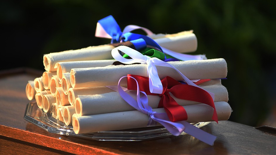 A pile of graduation diplomas on a wooden table, with white, red, blue, purple, and green ribbons tied around them.