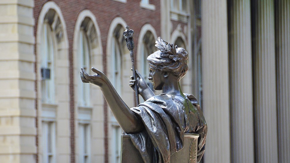 Statue of Alma Mater, a classically-robed woman sitting down with a laurel wreath in her hair and a scepter in her hand, view from the side. 