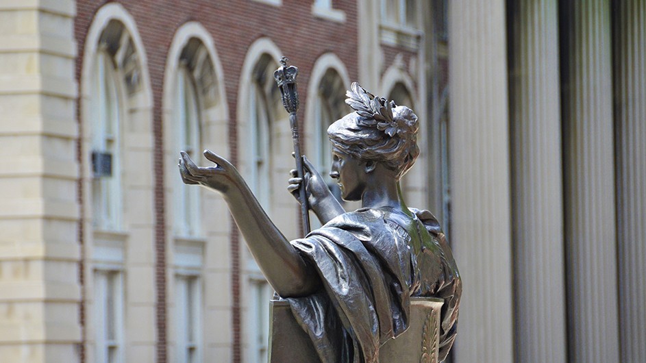 Eileen Barroso Statue of Alma Mater, a classically-robed woman sitting down with a laurel wreath in her hair and a scepter in her hand, view from the side.