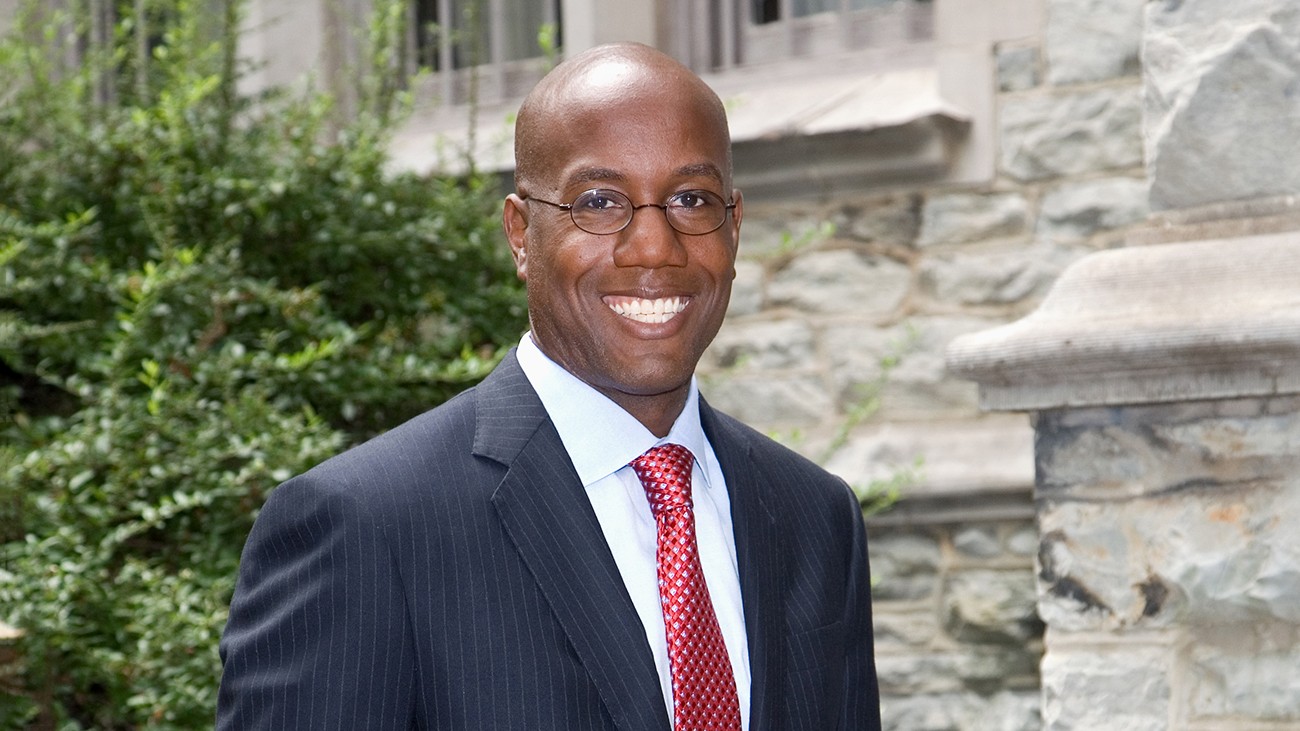Headshot of Jason Wingard, Dean of the School of Professional Studies, a dark skinned man with glasses and a clean shaven head in a dark blue suit, white shirt and red tie