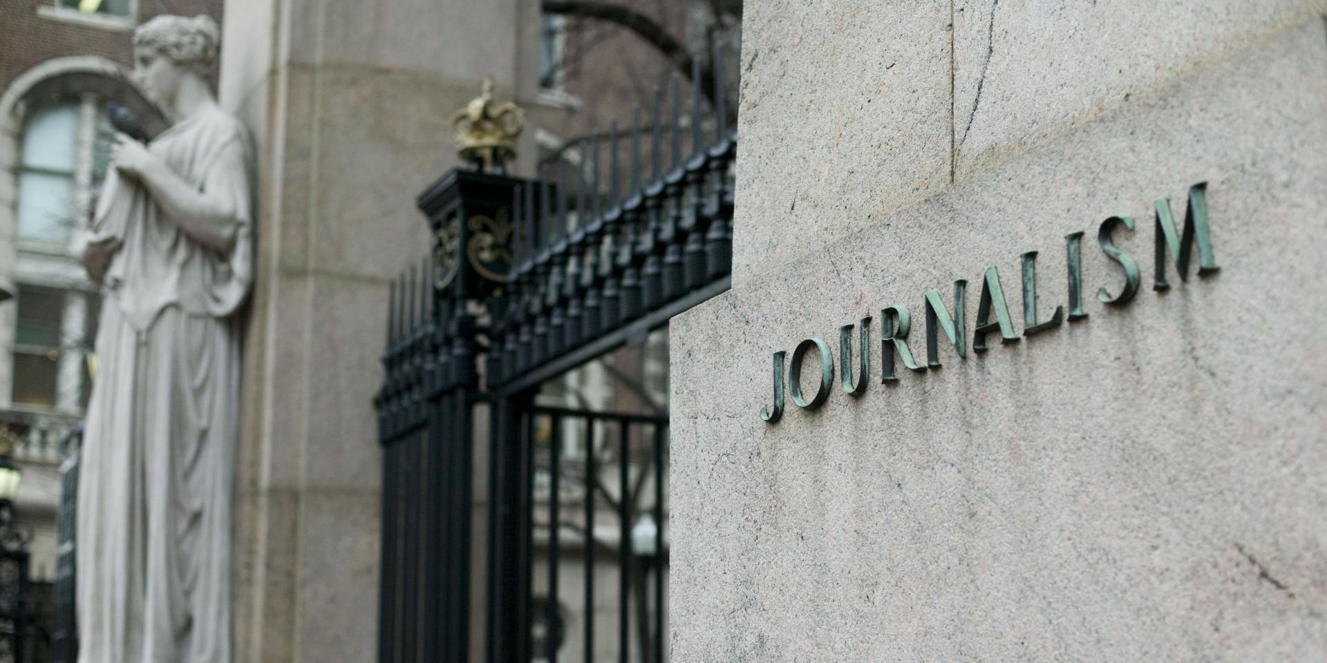 A close up view of the journalism building outside Columbia's Main Gate, with a statue in the background and the word "journalism" inscribed on the building in the foreground.