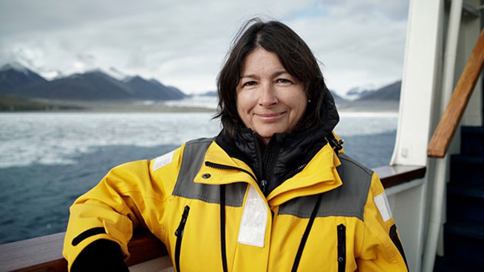 Headshot of a woman with dark hair, in a heavy yellow jacket, on a boat against an icy sea backdrop