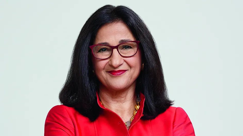 Minouche Shafik, a woman with dark brown hair and glasses, in a red top. 