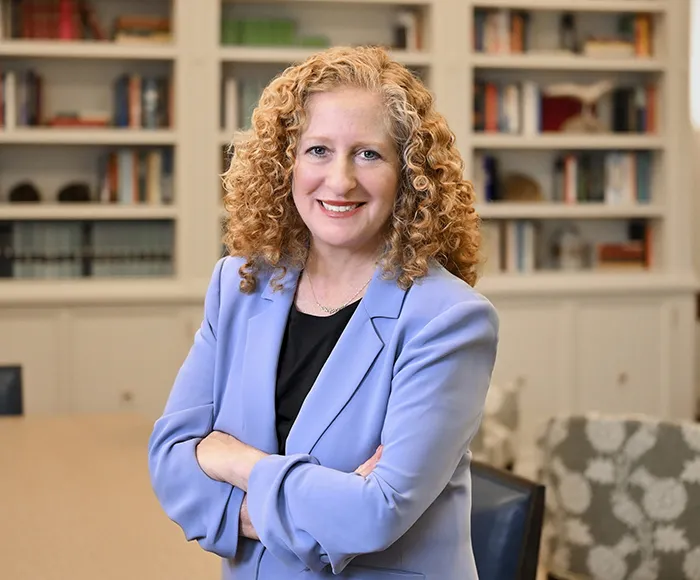 A woman with light hair in a blue suit standing in front of a bookcase.