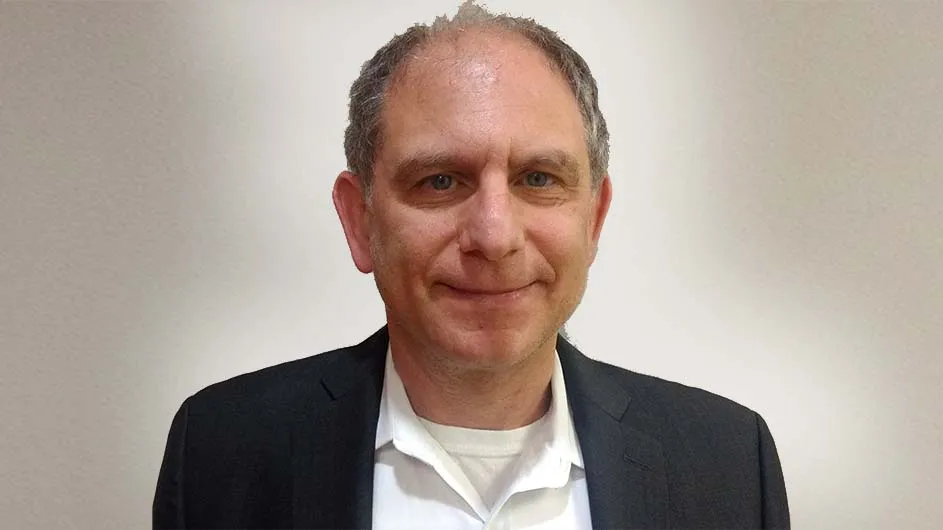 A man with short hair in a suit, against a light gray backdrop. 