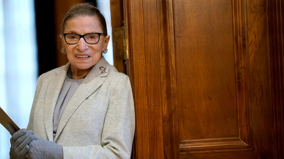 A woman with dark hair and glasses, in a light gray suit, against the backdrop of a medium-brown wood door. 