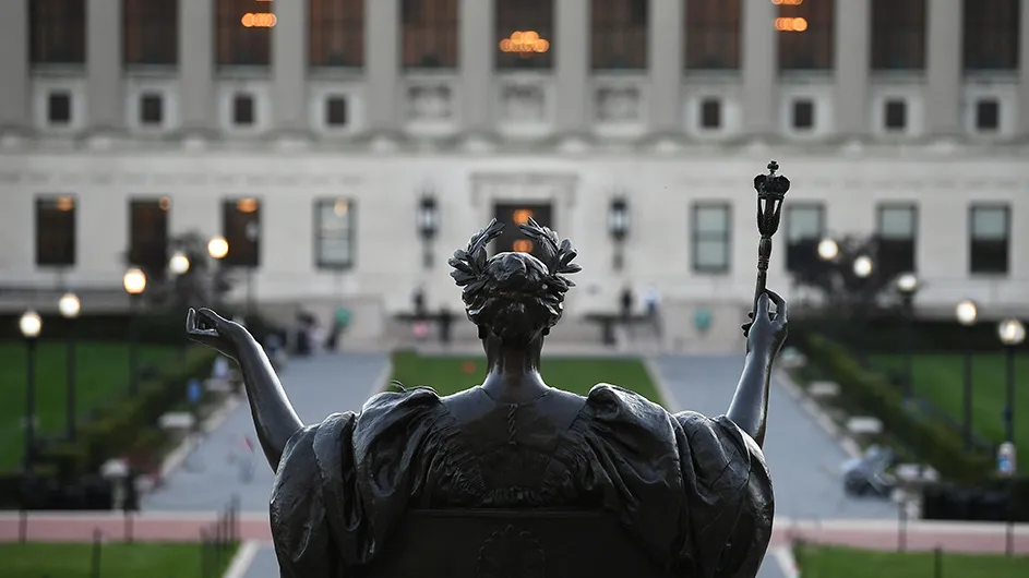 a view of the back of alma mater, a bronze statue of a woman with a laurel crown on her head and a scepter, in front of butler library, a classical building.