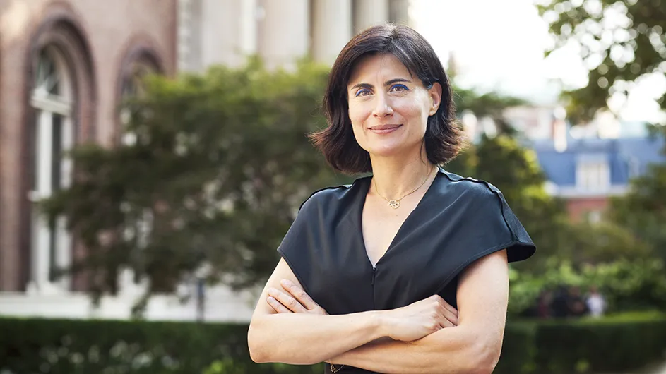 A woman with medium-length dark hair in a black top standing outside on Columbia's campus with her arms folded in front of her. 