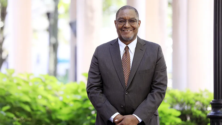 A man standing outside, in a grey suit and tie, smiling.