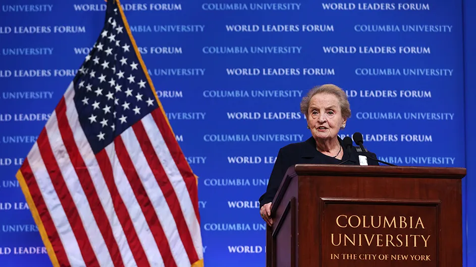 Madeleine Albright, a woman with short hair, standing at a podium in front of a blue backdrop and an American flag.
