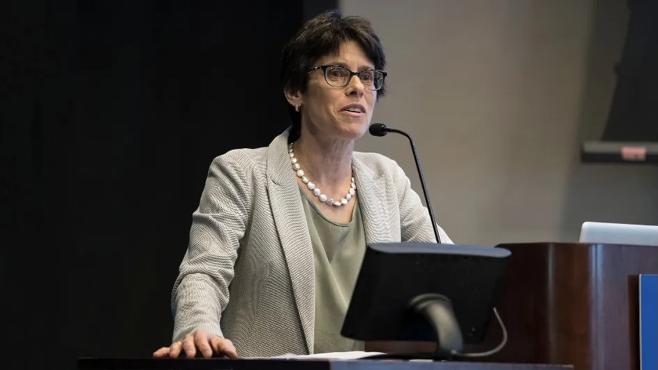 A woman with short, dark hair and glasses in a cream-colored suit, speaking at a lecturn. 