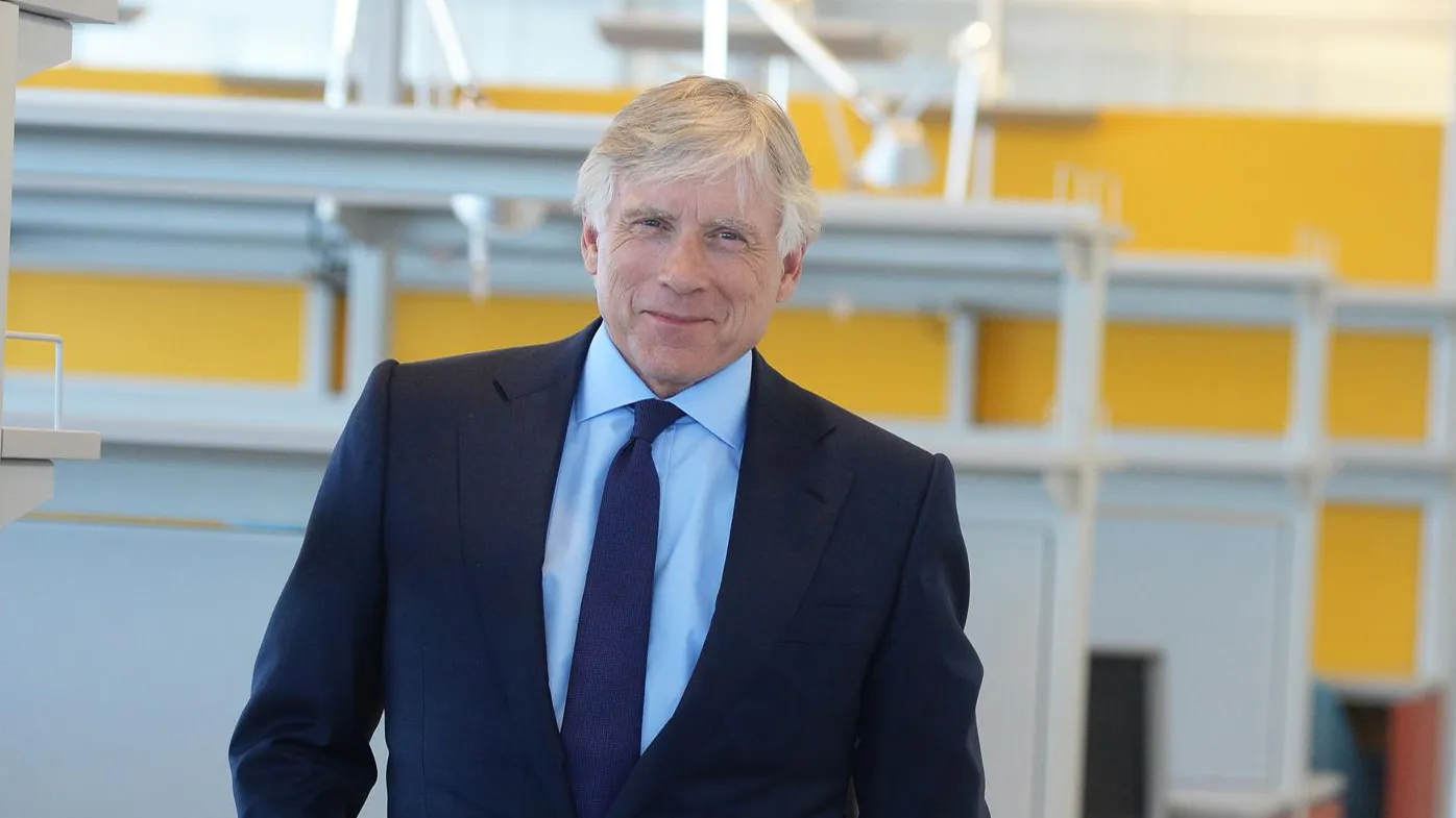 Man with white hair in a navy colored suit and tie. 