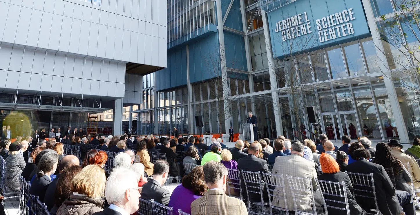A group of people are seated as President Lee Bollinger addresses the crowd at the opening of the Manhattanville campus. The two first buildings are in the background.