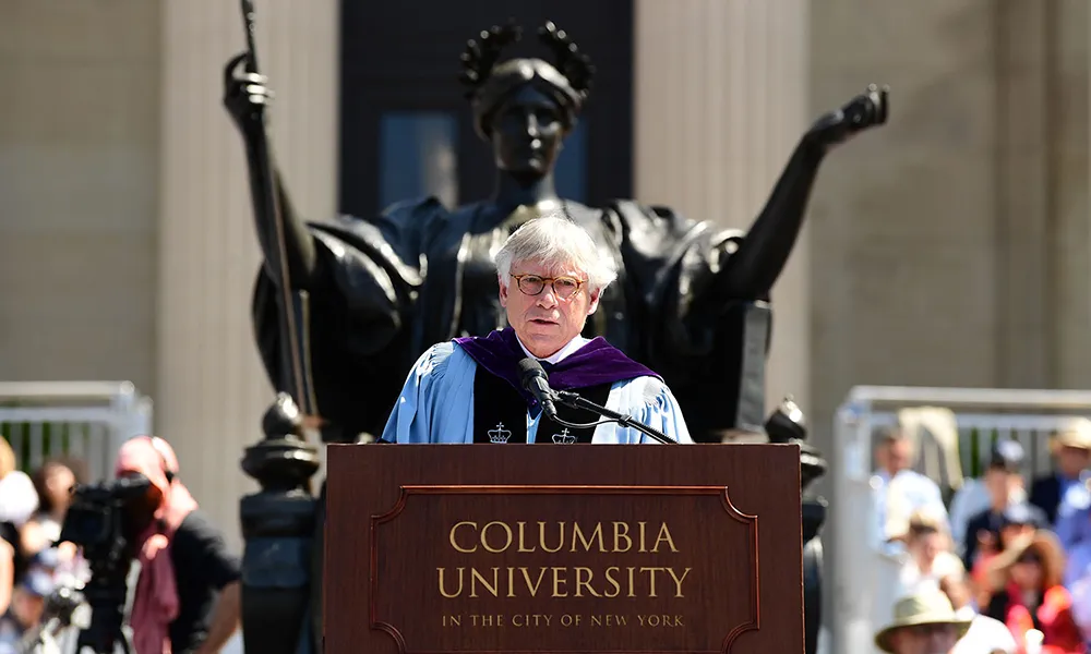 Photo of President Bollinger speaking at Commencement, at a lectern, in front of Columbia's statue of Alma Mater.