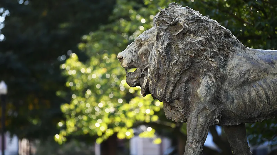 A gray stone statue of a lion, against a backdrop of green foliage. 